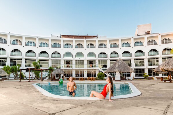 A pool area with a large building in the background, including a pool, lounge chairs, and umbrellas. - Uxmal Resort Maya (Muna)