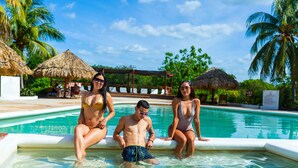 Área de piscina com água cristalina, palmeiras e guarda-sóis de palha.