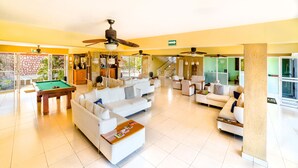 Uma sala de estar ampla com mesa de bilhar, vários sofás, ventiladores de teto e grandes janelas com vista para o exterior.