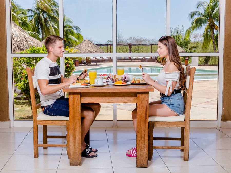 A dining table with chairs, a window with a view of a pool and palm trees, and a tropical setting.