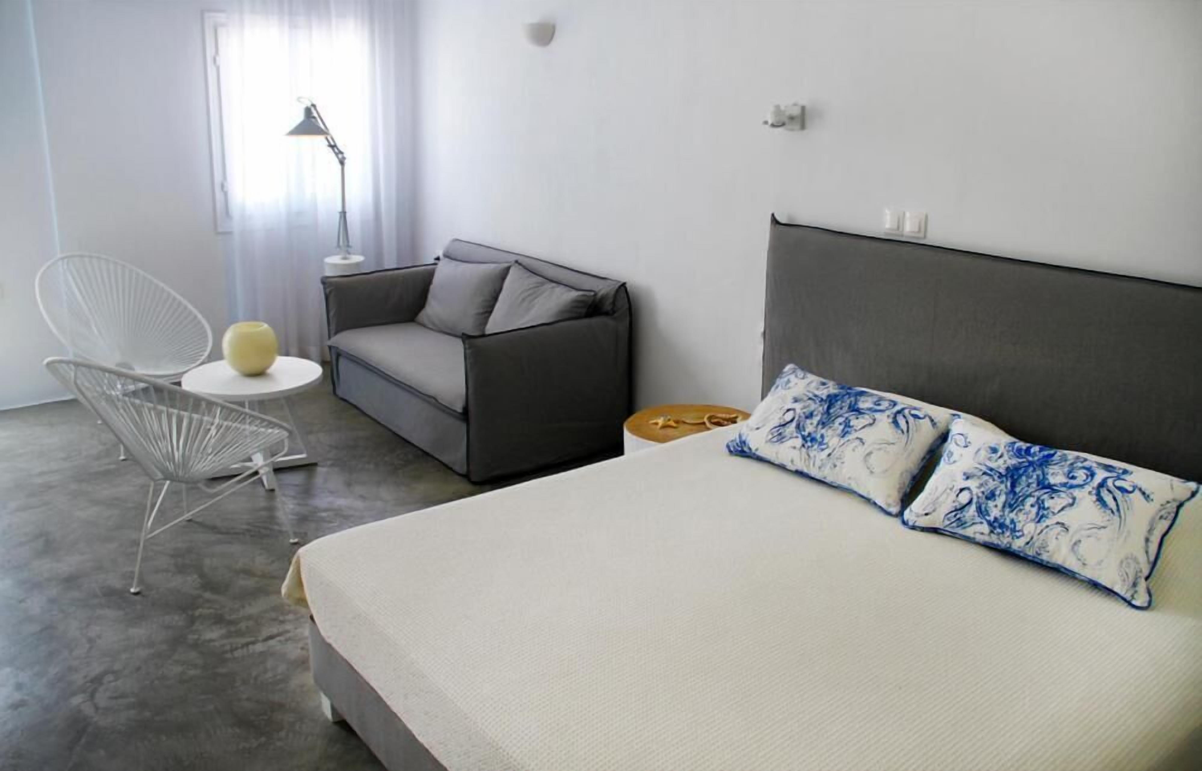 A modern bedroom with a grey sofa, a white chair, a round side table, and a bed with patterned pillows.