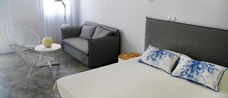 A modern bedroom with a grey sofa, a white chair, a round side table, and a bed with patterned pillows.