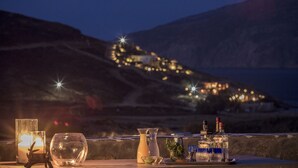 A table with a candle, a glass pitcher, and bottles, set against a night view of a coastal town with lights. - Anamar Mykonos Natural Retreat (Mykonos)