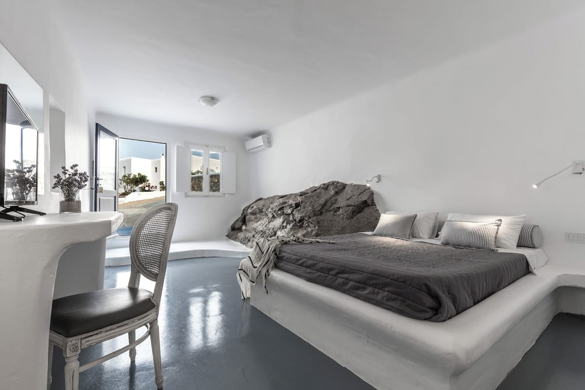 A modern bedroom with a large bed, a desk, and a chair. There is a view of the outdoors through large windows.