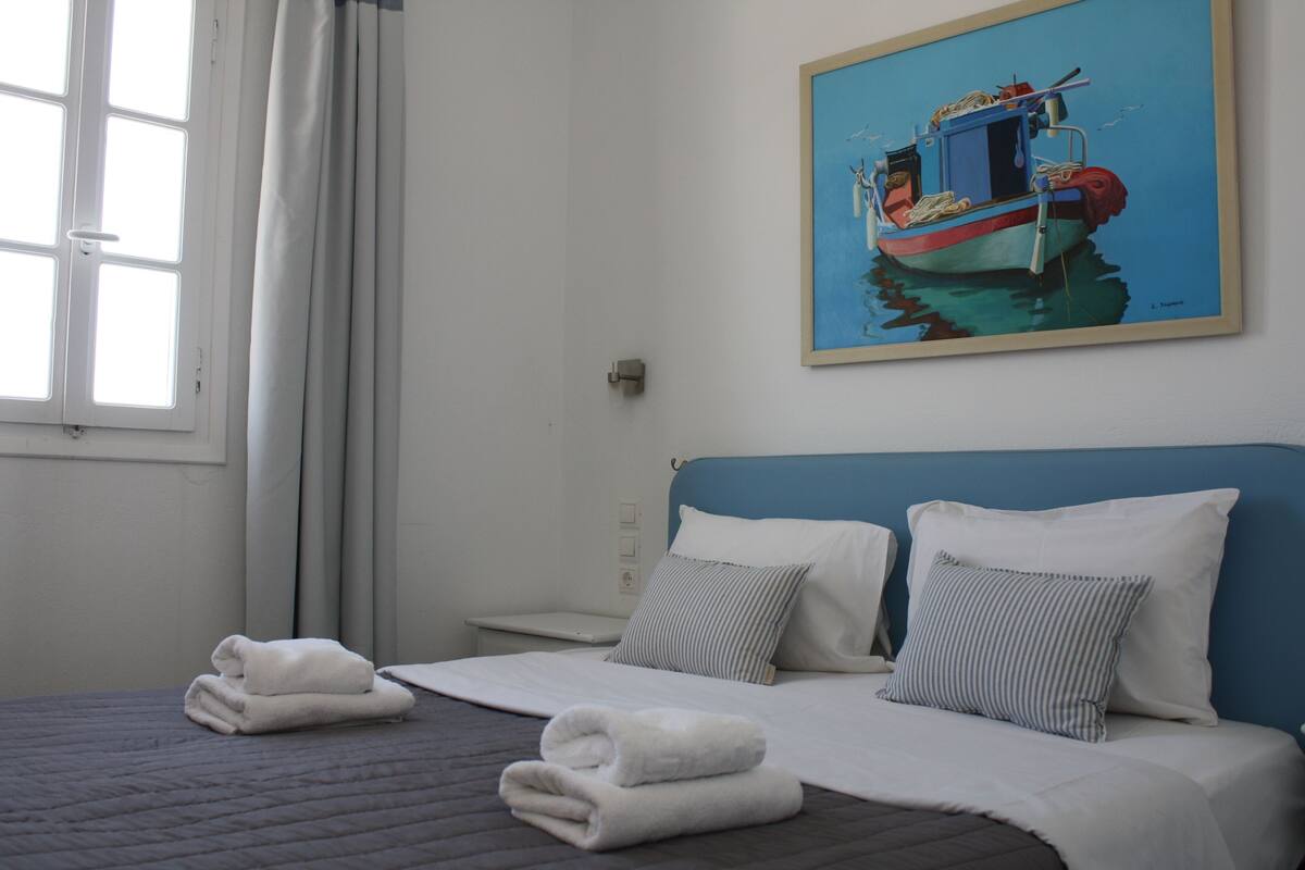 A neatly made bed with white linens and two pillows, a painting of a boat on the wall, and a window with curtains.