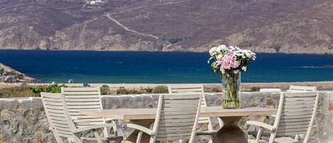 A table with chairs and a vase of flowers, overlooking a coastal landscape with mountains and the sea.