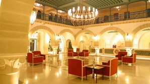 Un vestíbulo amplio con techo alto, candelabros ornamentales y zonas de estar con asientos rojos.