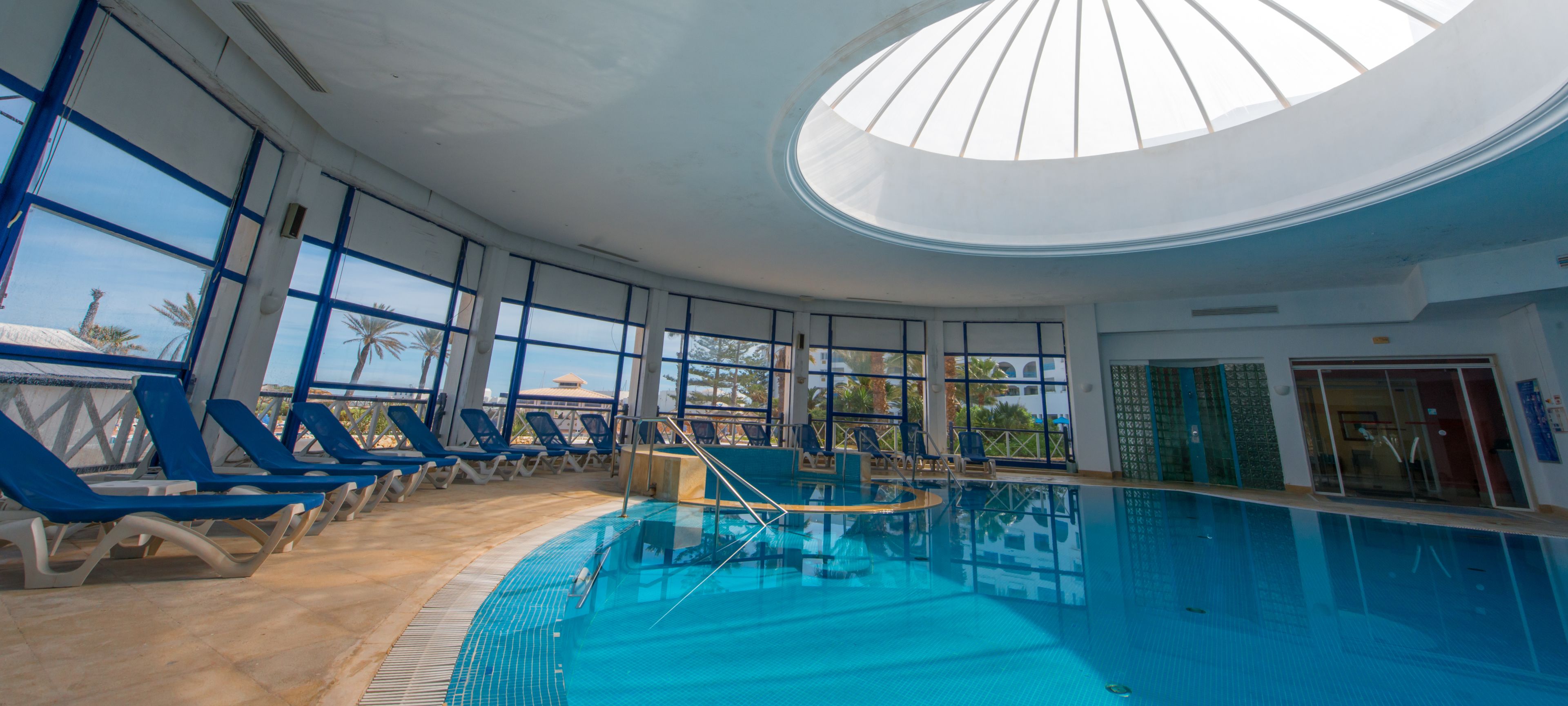 A large indoor swimming pool with blue water, surrounded by lounge chairs and large windows.