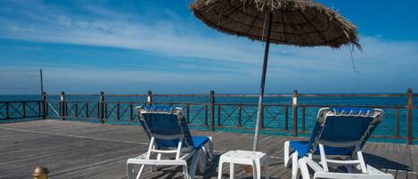 A wooden deck with two white lounge chairs and a thatched umbrella, overlooking a clear blue sea.