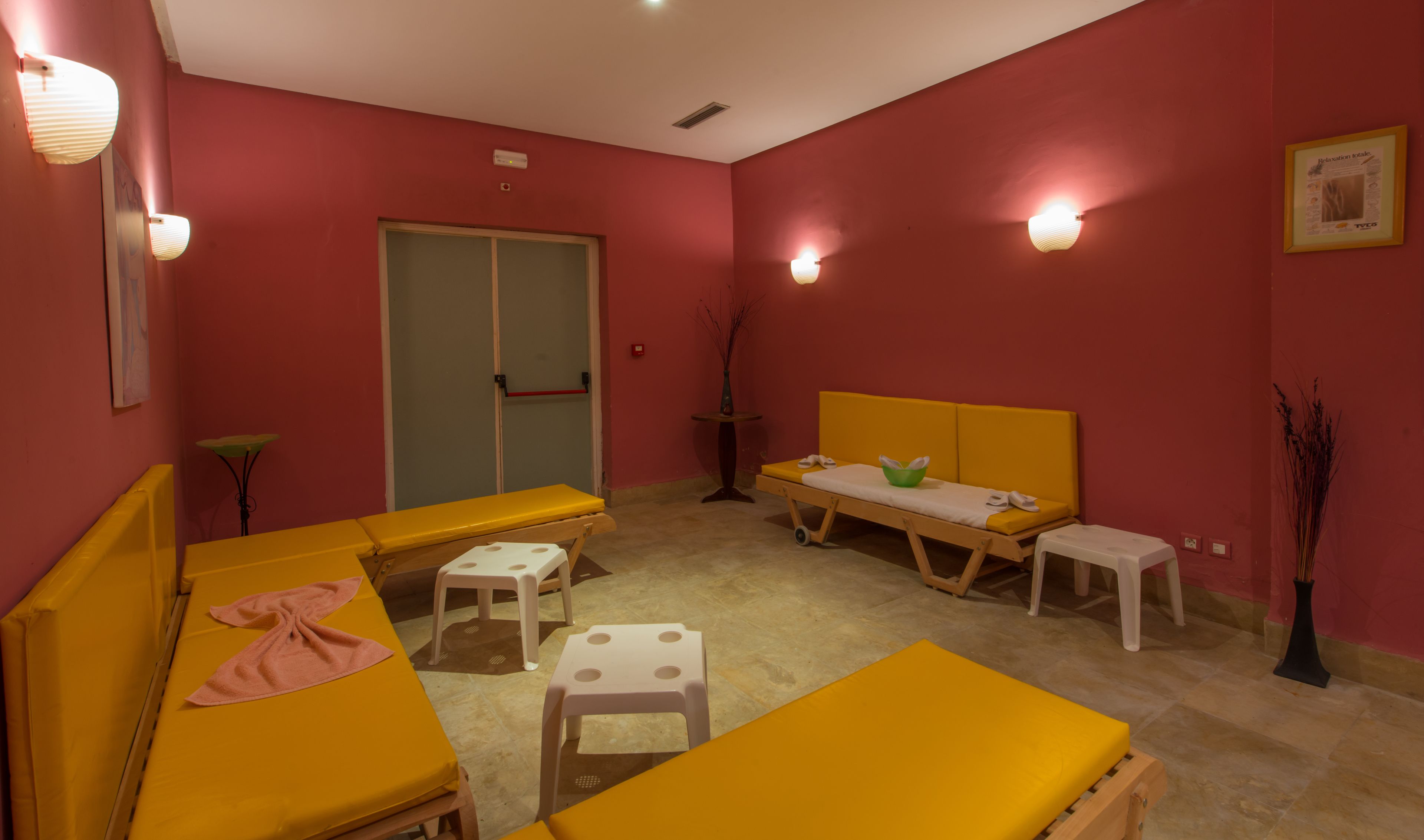 A room with yellow beds, a small table, and a bench. There are wall-mounted lights and a door with a glass panel.