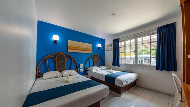 Quarto com duas camas, uma parede azul decorativa, uma janela com cortinas e uma luminária no teto.