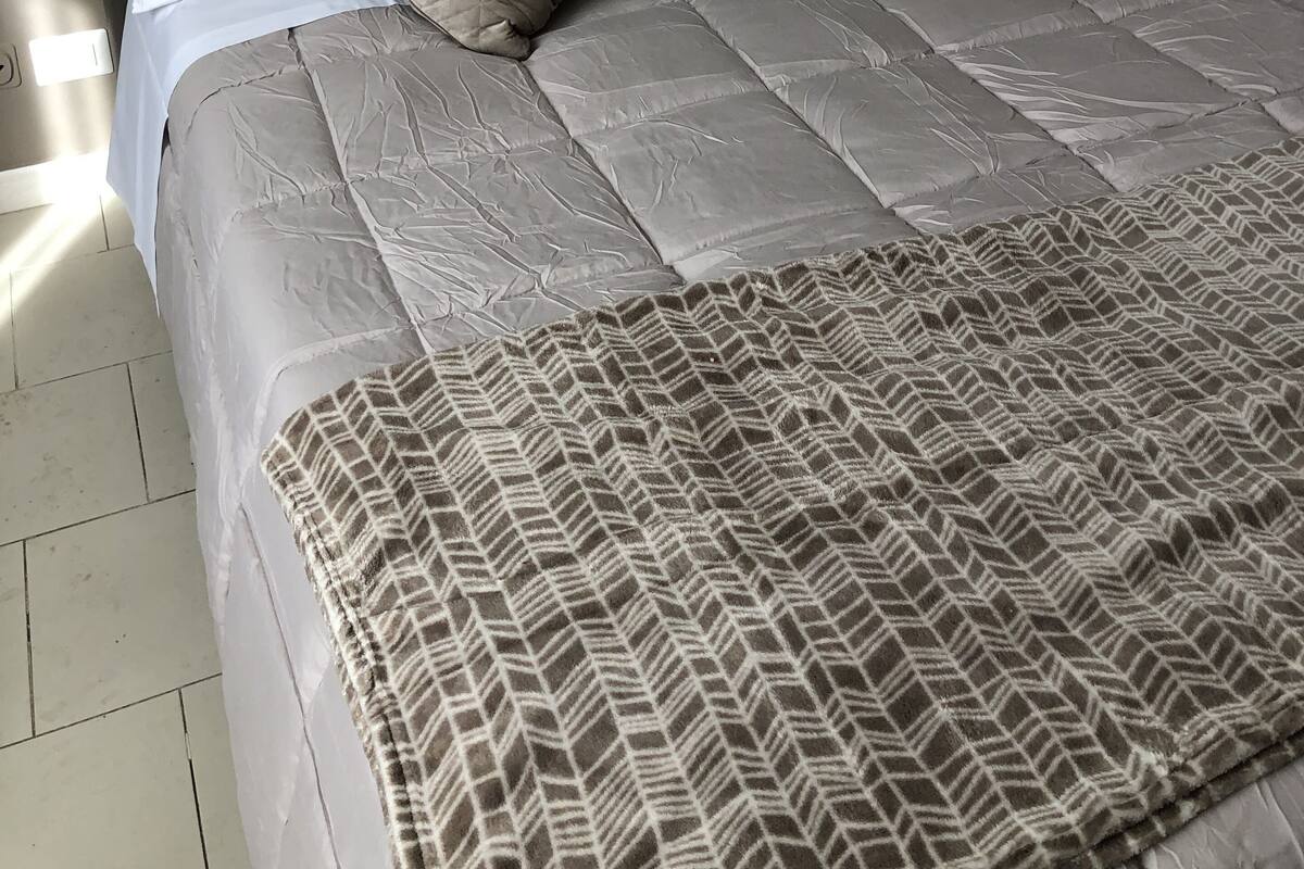 A neatly made bed with a patterned bedspread and two pillows, a bedside lamp, and a wall socket.
