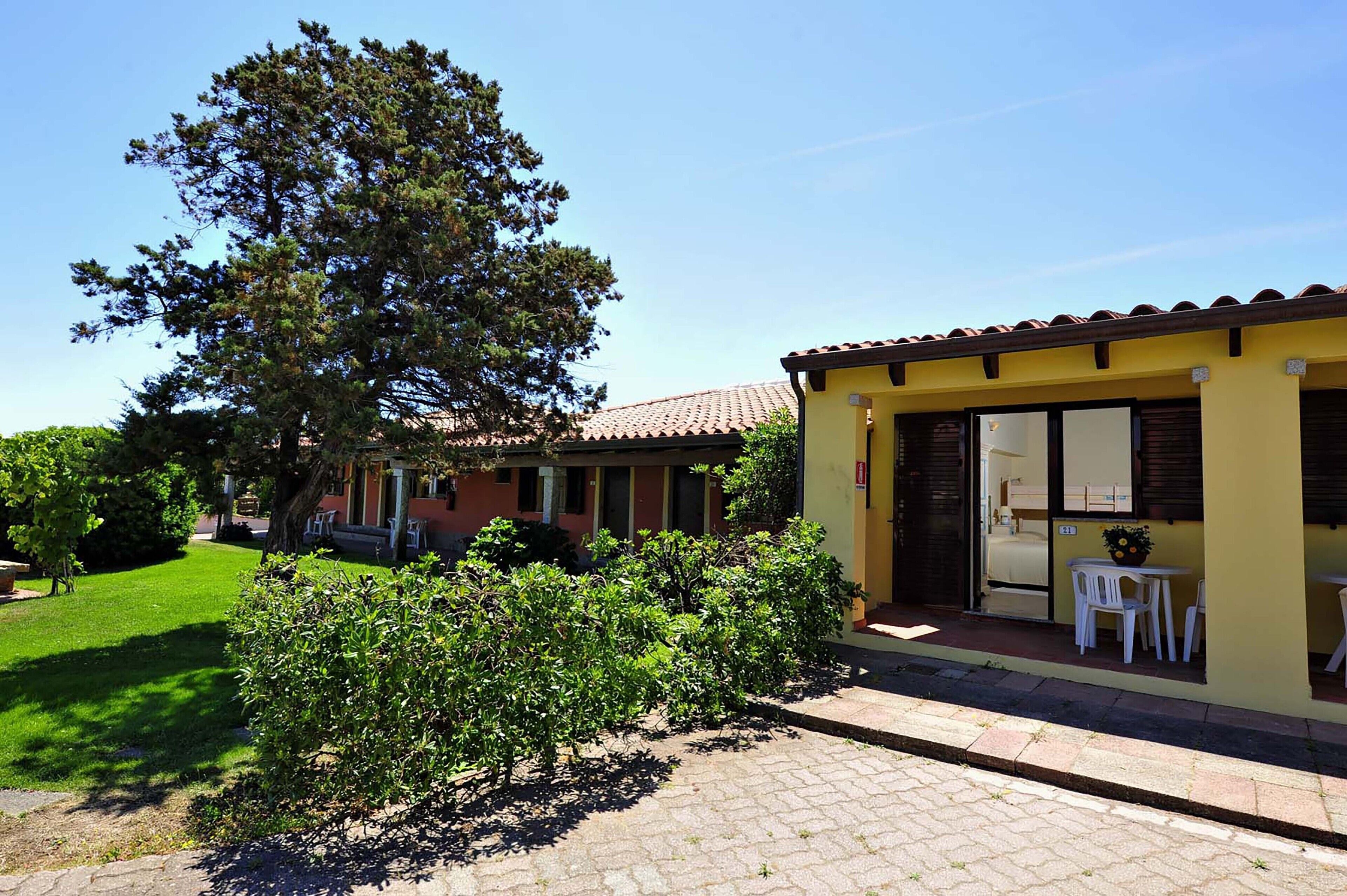 A yellow building with a tiled roof, a small patio with white chairs, and a large tree in the background.