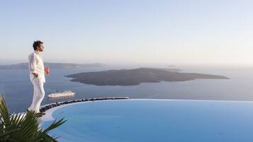 Um homem de camisa e calças brancas parado perto de uma piscina, com vista para o mar e um navio ao longe.