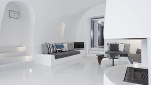A modern, minimalist living room with a white color scheme, a glass coffee table, and a built-in fireplace. - Alta Vista Suites (Santorini)