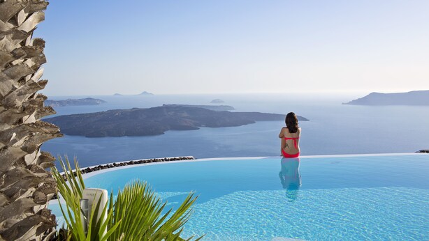 A woman in a red swimsuit standing at the edge of a infinity pool with a view of the sea and islands. - Alta Vista Suites (Santorini)