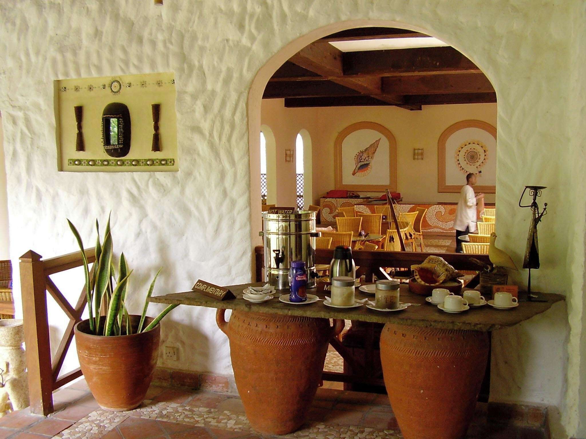 A rustic interior with a stone wall, a wooden table with tea sets, a potted plant, and a metal sculpture.