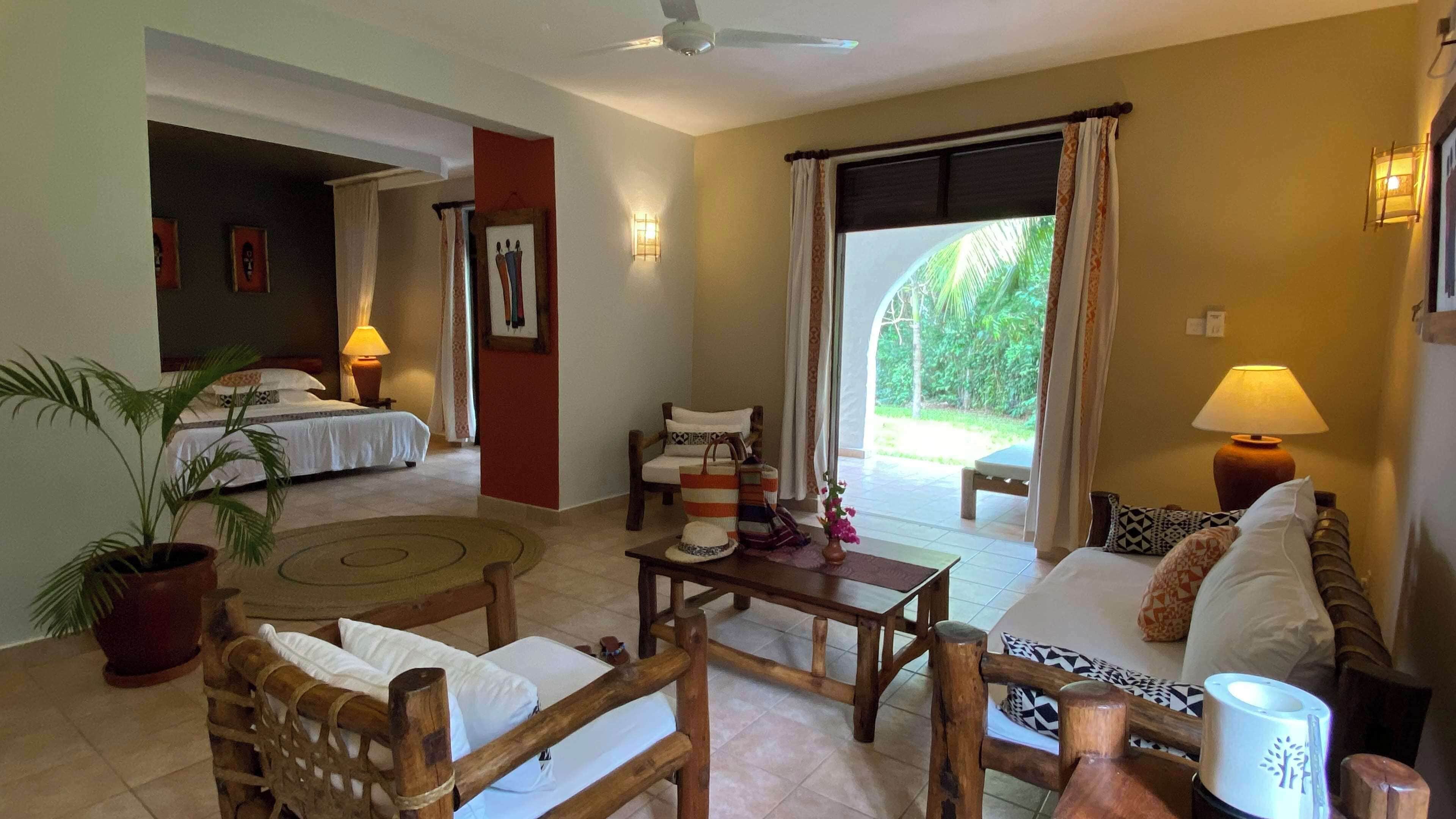 A hotel room with a bed, a sitting area with a table and chairs, a potted plant, and a view of the outdoors.