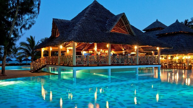 Un resort con estructuras de techo de paja, una piscina con azulejos azules y un puente de madera.