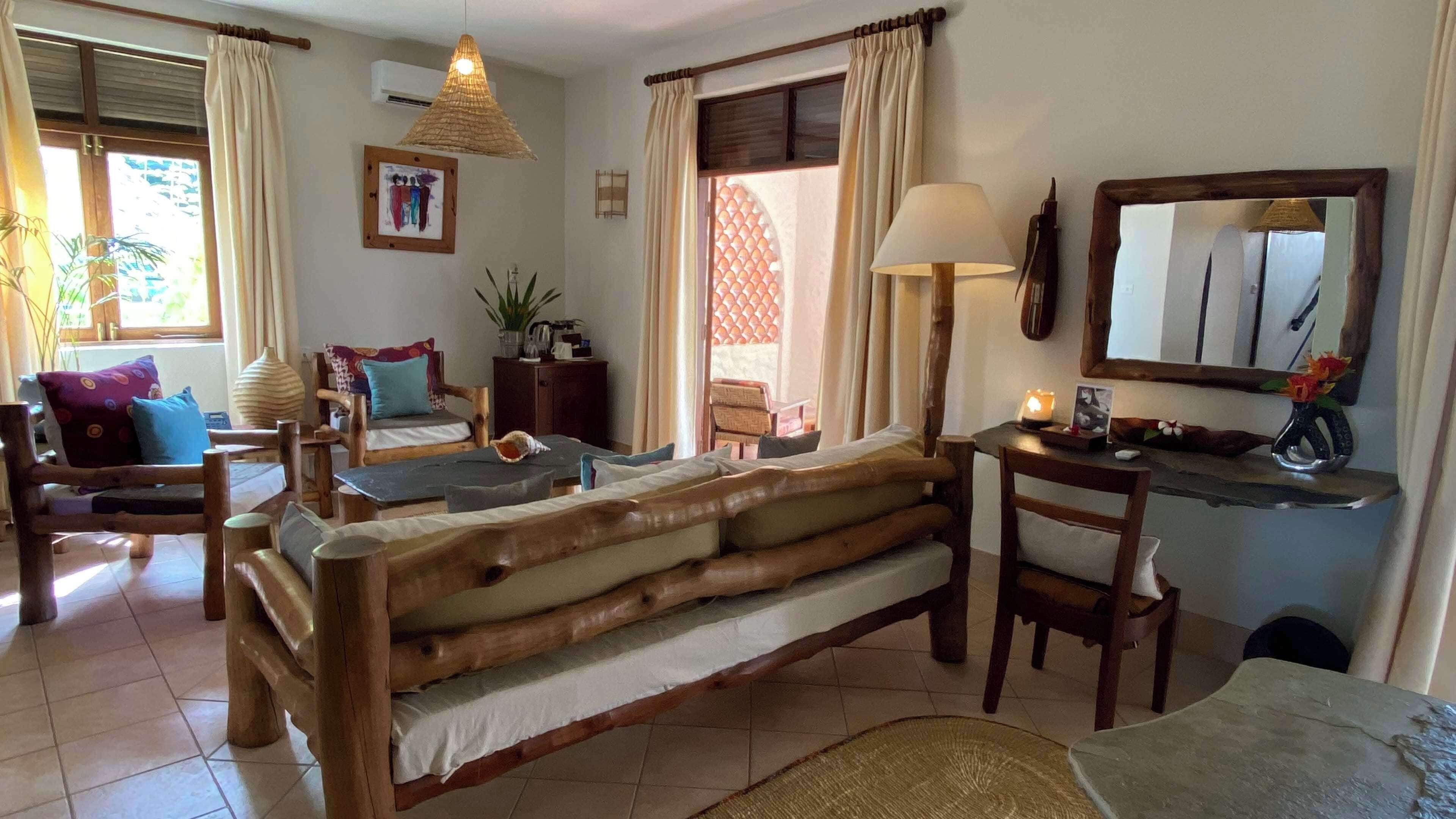 A cozy living room with a wooden sofa, a dining table, and a chair. There is a lamp, a mirror, and a potted plant.