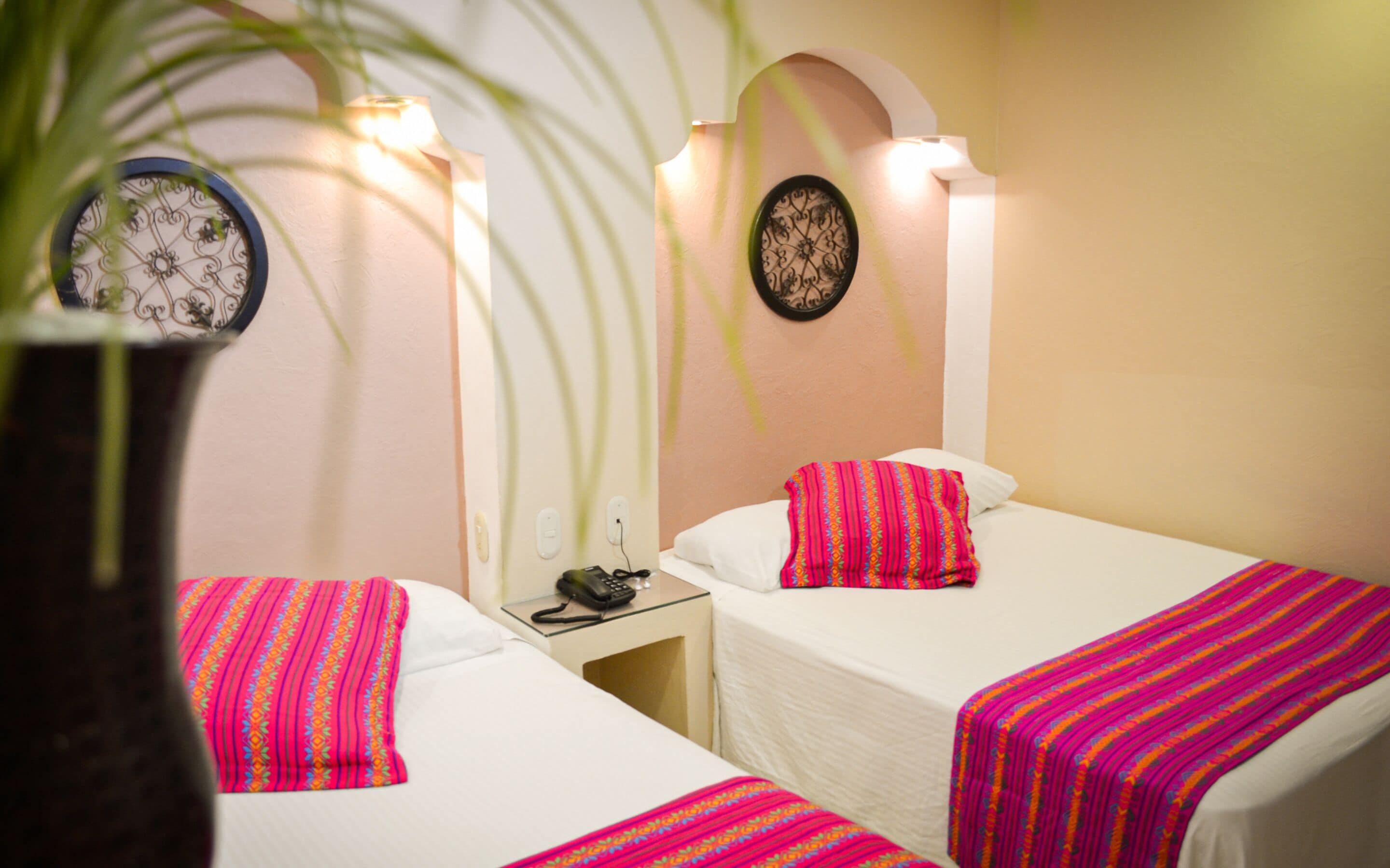 Habitación de hotel con dos camas, cada una con una colcha estampada de color rosa. Hay relojes de pared y una mesita con un teléfono y una lámpara.