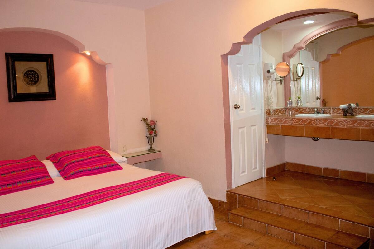 A bedroom with a bed, a nightstand, a vase with flowers, and a bathroom with a sink and mirror.