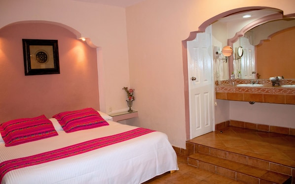 Un dormitorio con cama, mesita de noche, un florero con flores y un baño con lavabo y espejo.