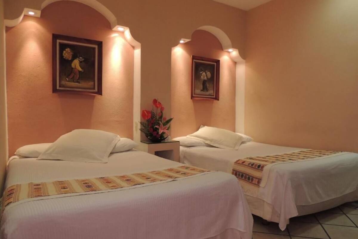 A hotel room with two beds, each with white linens and a patterned bedspread. There are two framed pictures on the walls, and a vase with flowers on the nightstand.