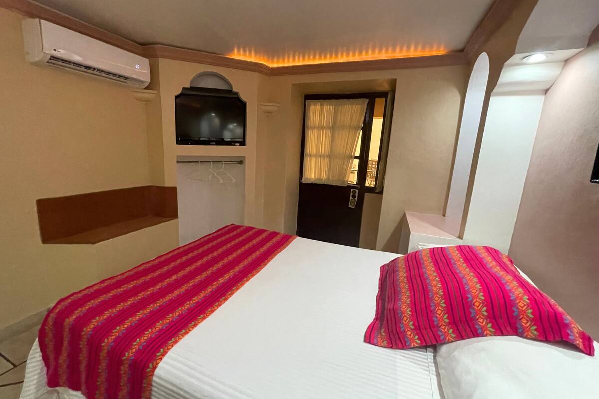 A hotel room with a bed, a TV, an air conditioner, and a door with a curtain.