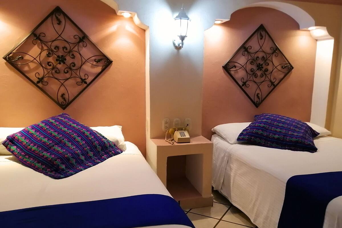 A hotel room with two beds, each with a patterned pillow and a blue bedspread. There is a bedside table with a telephone and a wall-mounted light fixture.