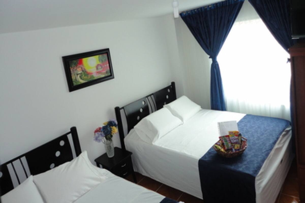 Habitación de hotel con dos camas individuales, una mesita de noche con un florero y un cuadro en la pared, además de una ventana con cortinas azules.