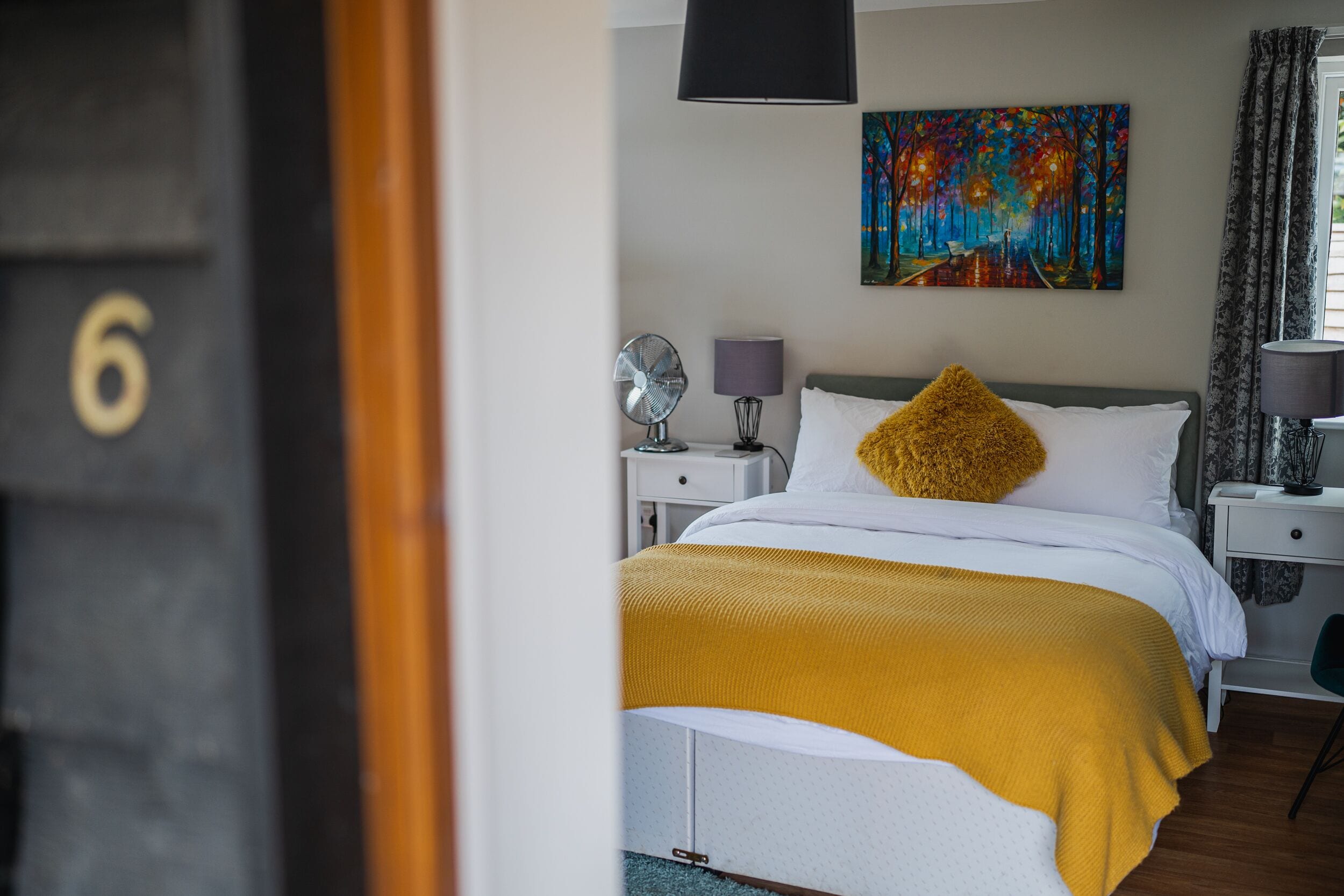 A bedroom with a bed, a nightstand, a lamp, a painting on the wall, and a window with curtains.