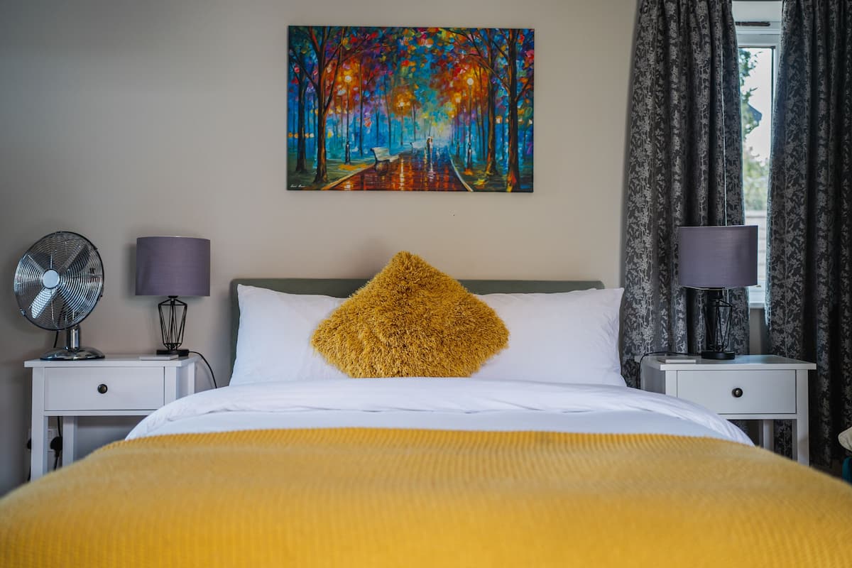 A hotel room with a bed, a nightstand with a lamp, a wall-mounted painting, and a window with curtains.