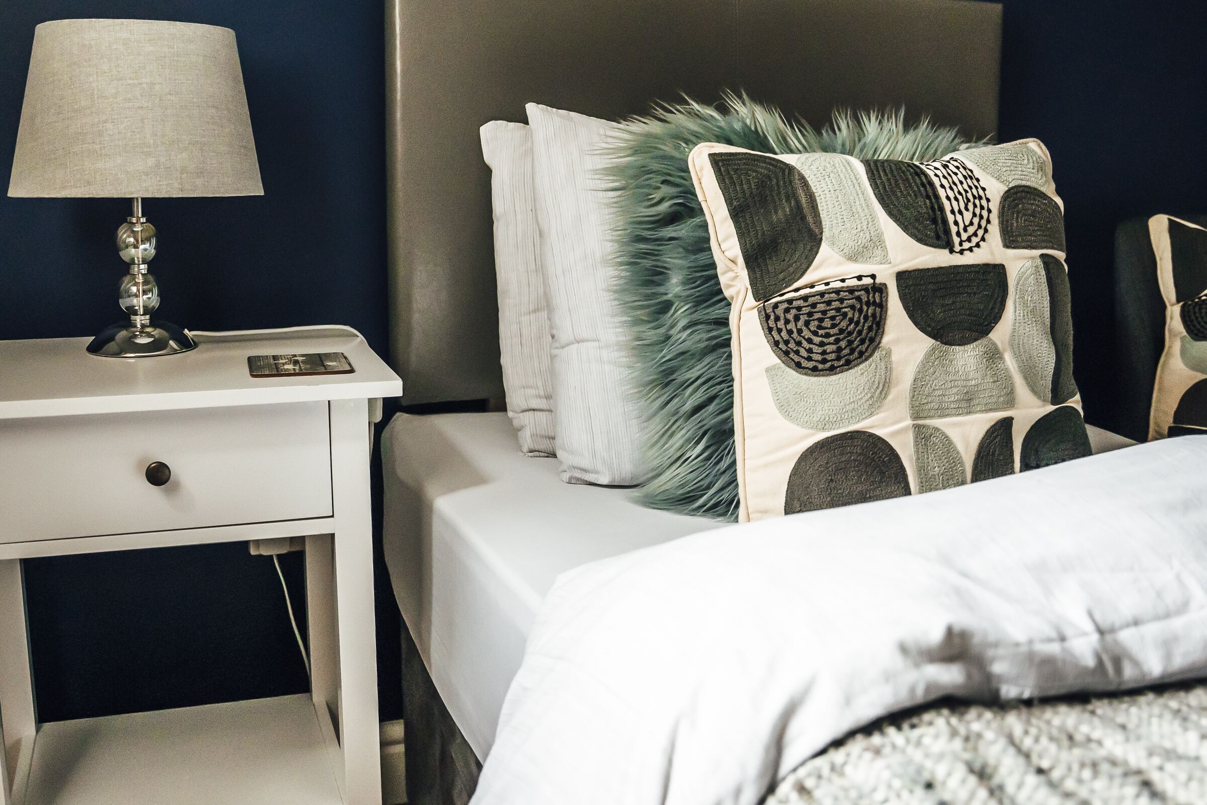 Een net opgemaakt bed met gestreepte kussens, een bedlampje en een witte nachtkastje met een lade.