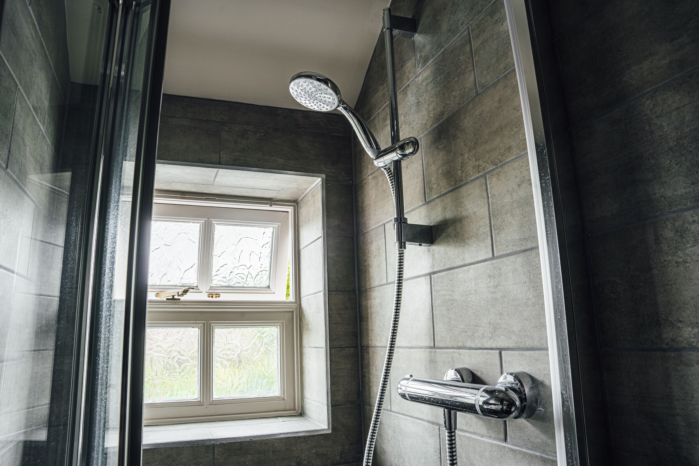 A shower with a handheld showerhead, a window, and a tiled wall.