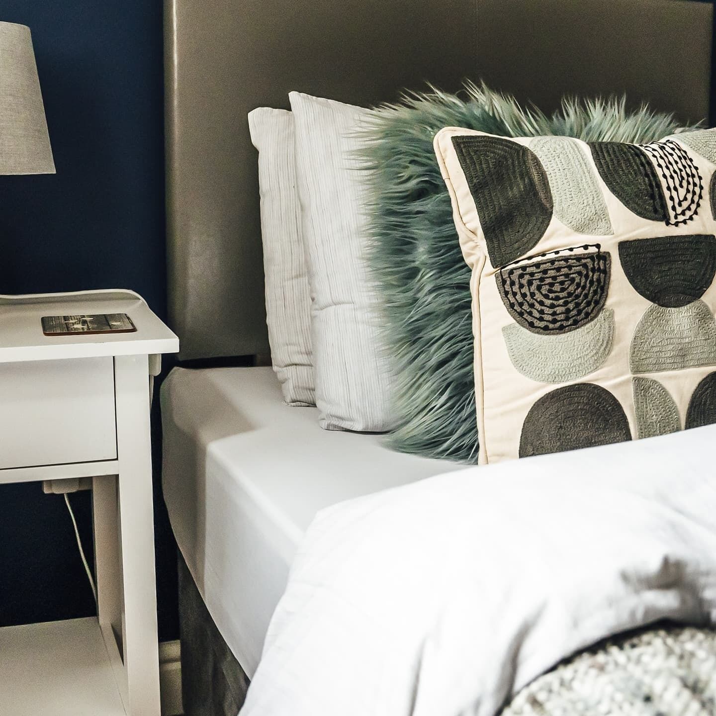 A neatly made bed with a patterned pillow and a white bedside table with a lamp.