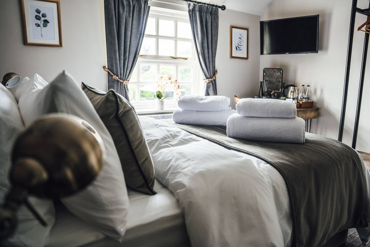 A neatly made bed with pillows and a stack of towels, a bedside table with a lamp, a window with curtains, and a flat-screen TV on the wall.