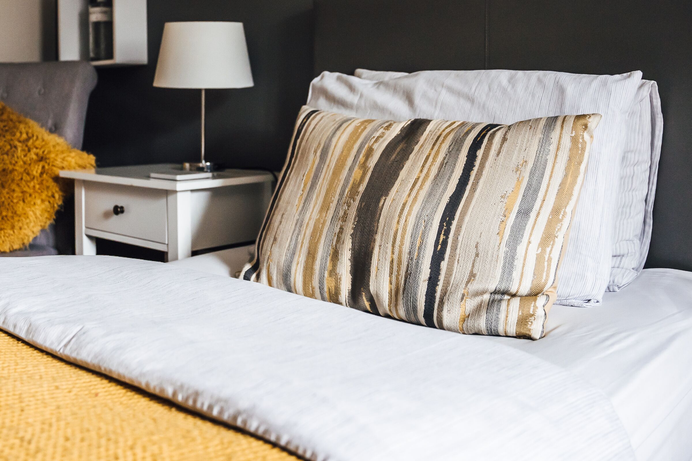 A neatly made bed with white linens and striped pillows, a bedside table with a lamp, and a dark wall in the background.
