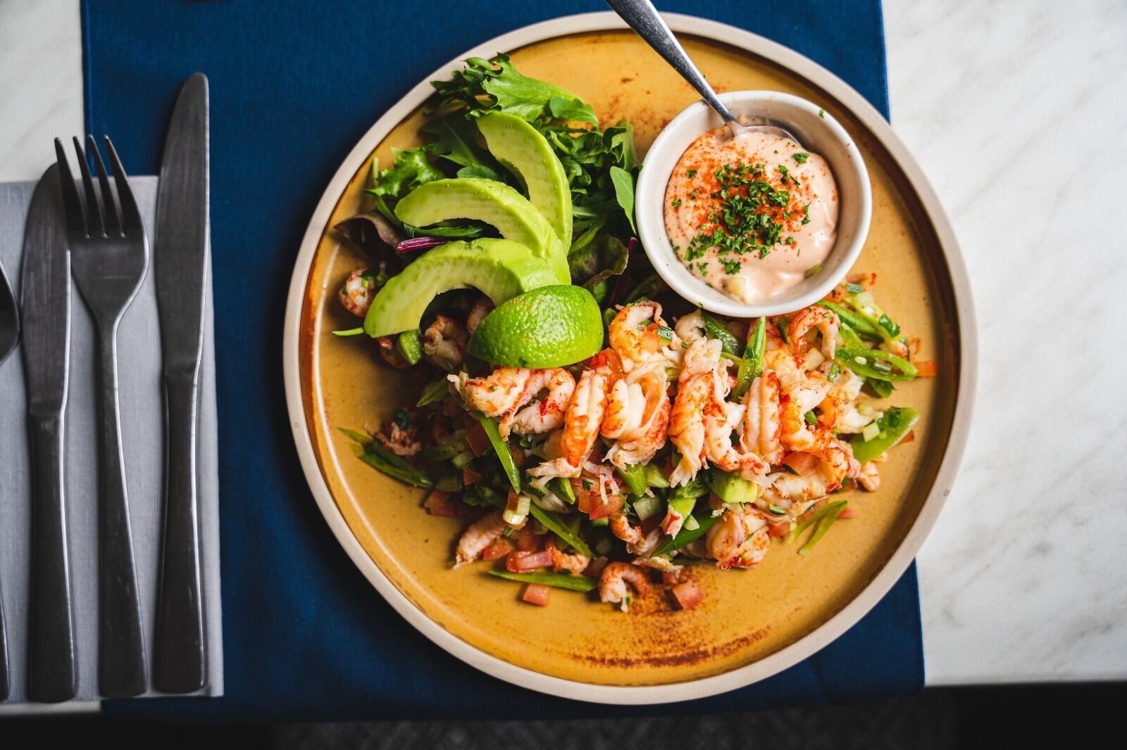 A plate of shrimp salad with avocado, lime, and a side of sauce, accompanied by a fork and knife on a blue napkin.