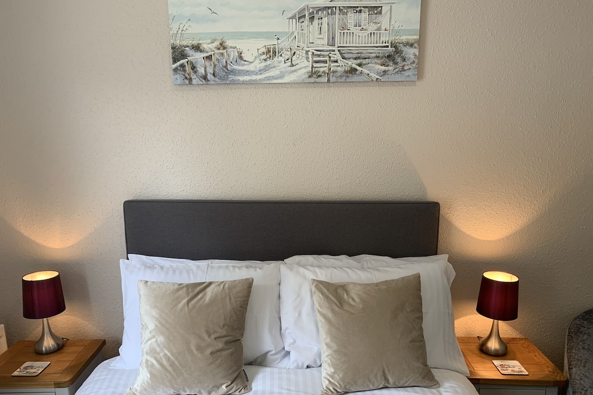A neatly made bed with white and beige bedding, two bedside lamps, and a painting of a beach scene above the bed.