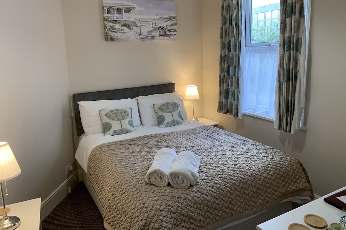 A neatly made bed with a quilted cover, two pillows with a floral design, a rolled towel, a bedside lamp, and a painting of a coastal scene on the wall.