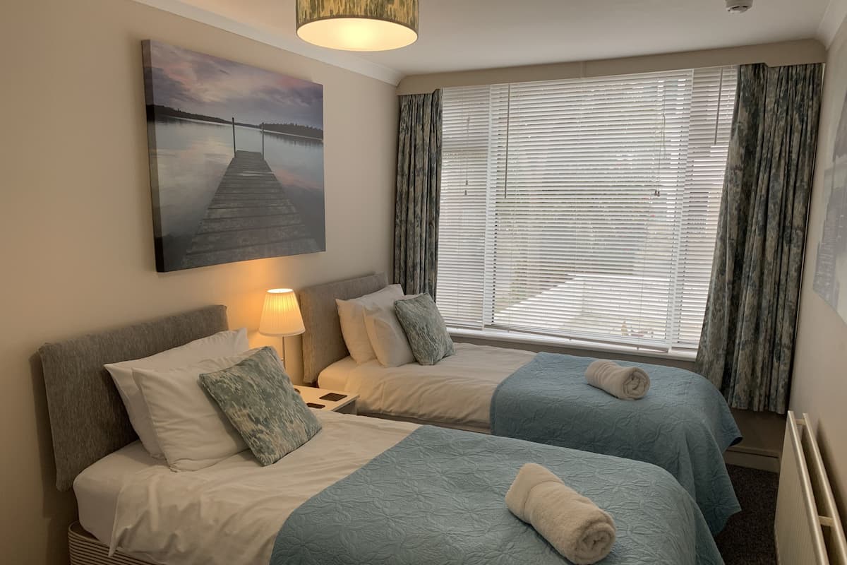 A hotel room with two beds, a nightstand, a lamp, a painting of a dock, and a window with curtains.