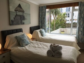 A hotel room with two beds, a painting of a beach scene, a window with a view of palm trees and buildings, and a set of towels on one bed. - The Chestnuts Guest House (Shanklin)