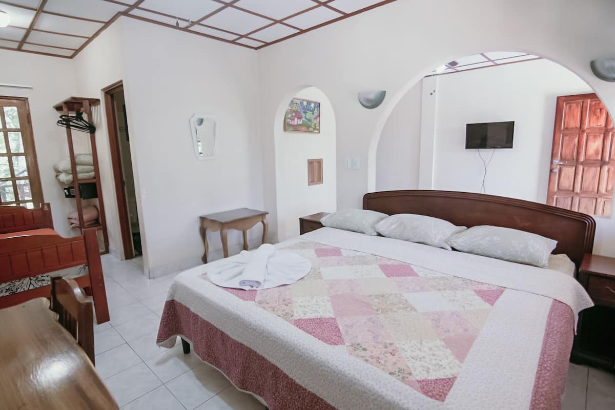 A bedroom with a bed, a wooden chair, a small table, and a TV mounted on the wall.