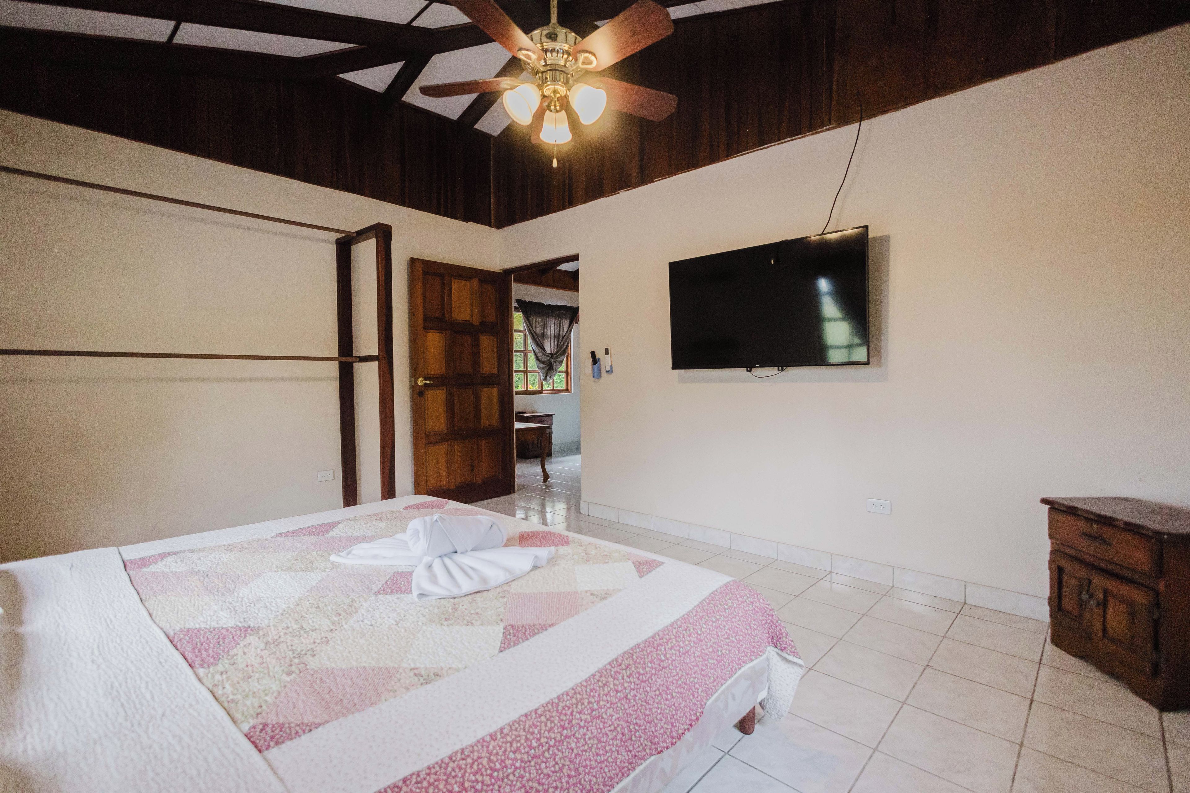 A bedroom with a bed, a ceiling fan, a TV mounted on the wall, and a wooden door.