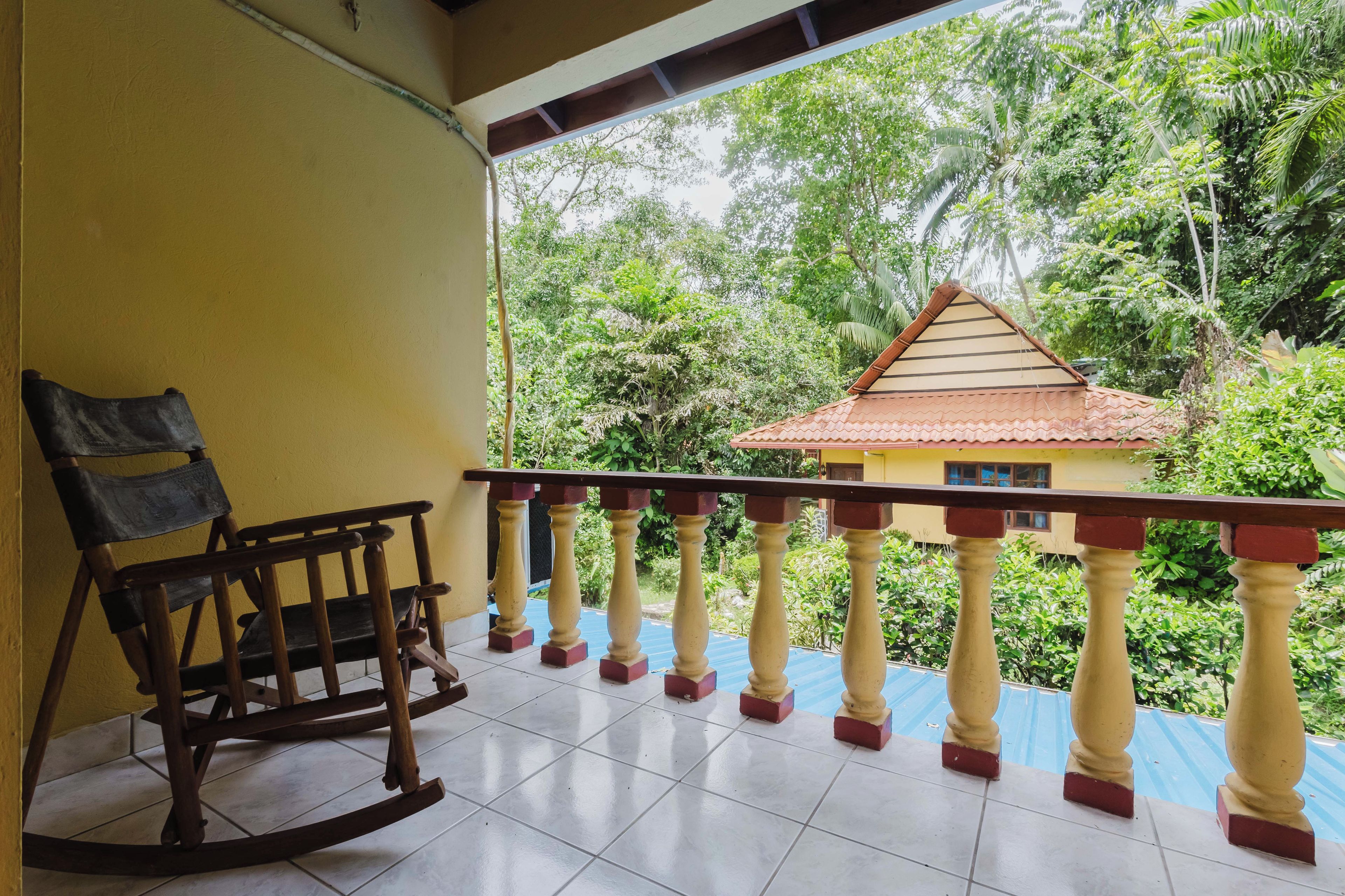 A balcony with a rocking chair, a tiled floor, and a view of a house surrounded by greenery.