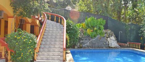A swimming pool with a tiled border, a staircase with a patterned railing, and a stone wall with greenery.