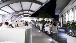 Un área de comedor al aire libre con mesas y sillas blancas, techo curvo y plantas en macetas.