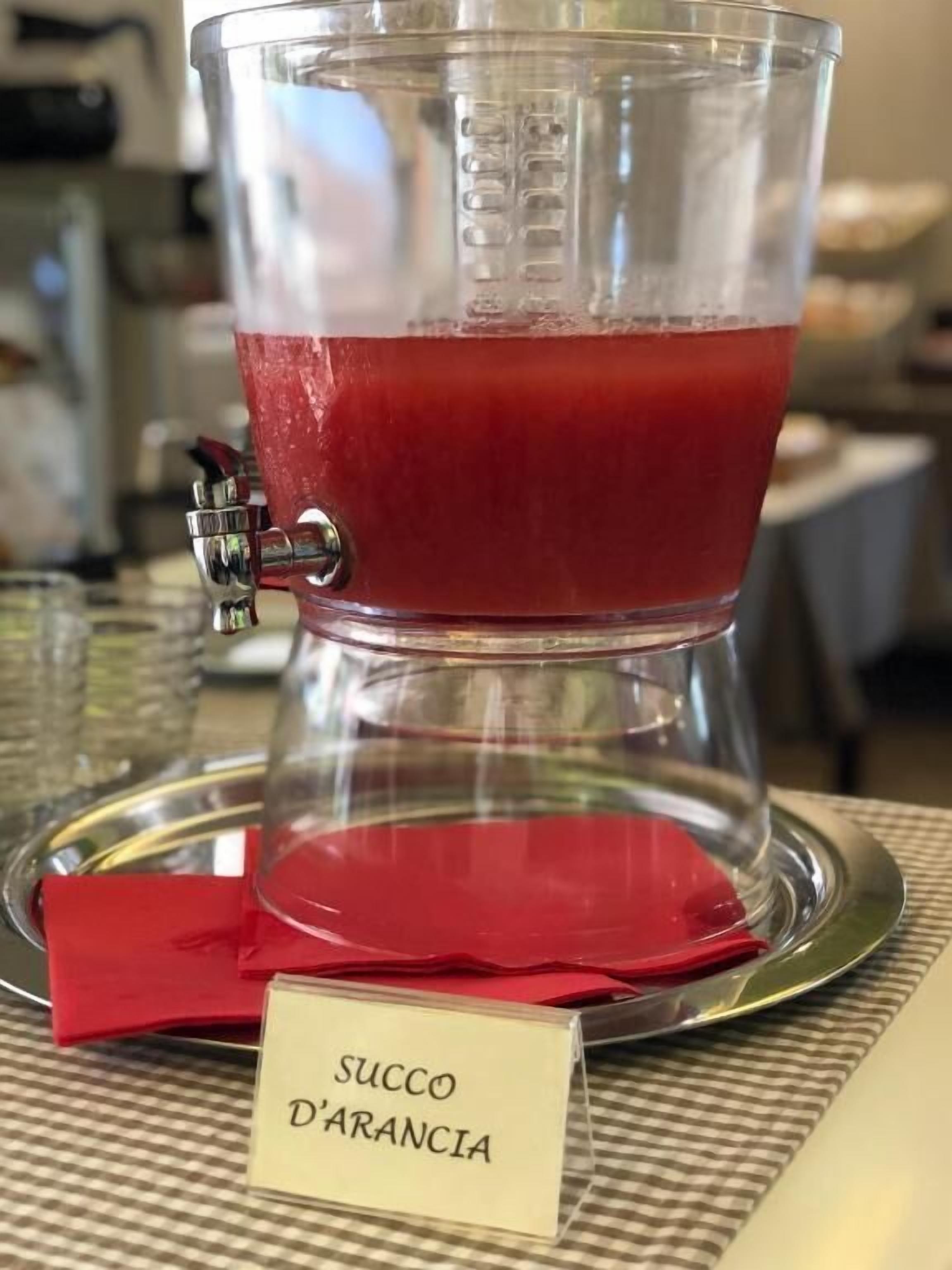 A clear glass cup with a red beverage, labeled "SUCCO D'ARANCIA," on a checkered tablecloth.