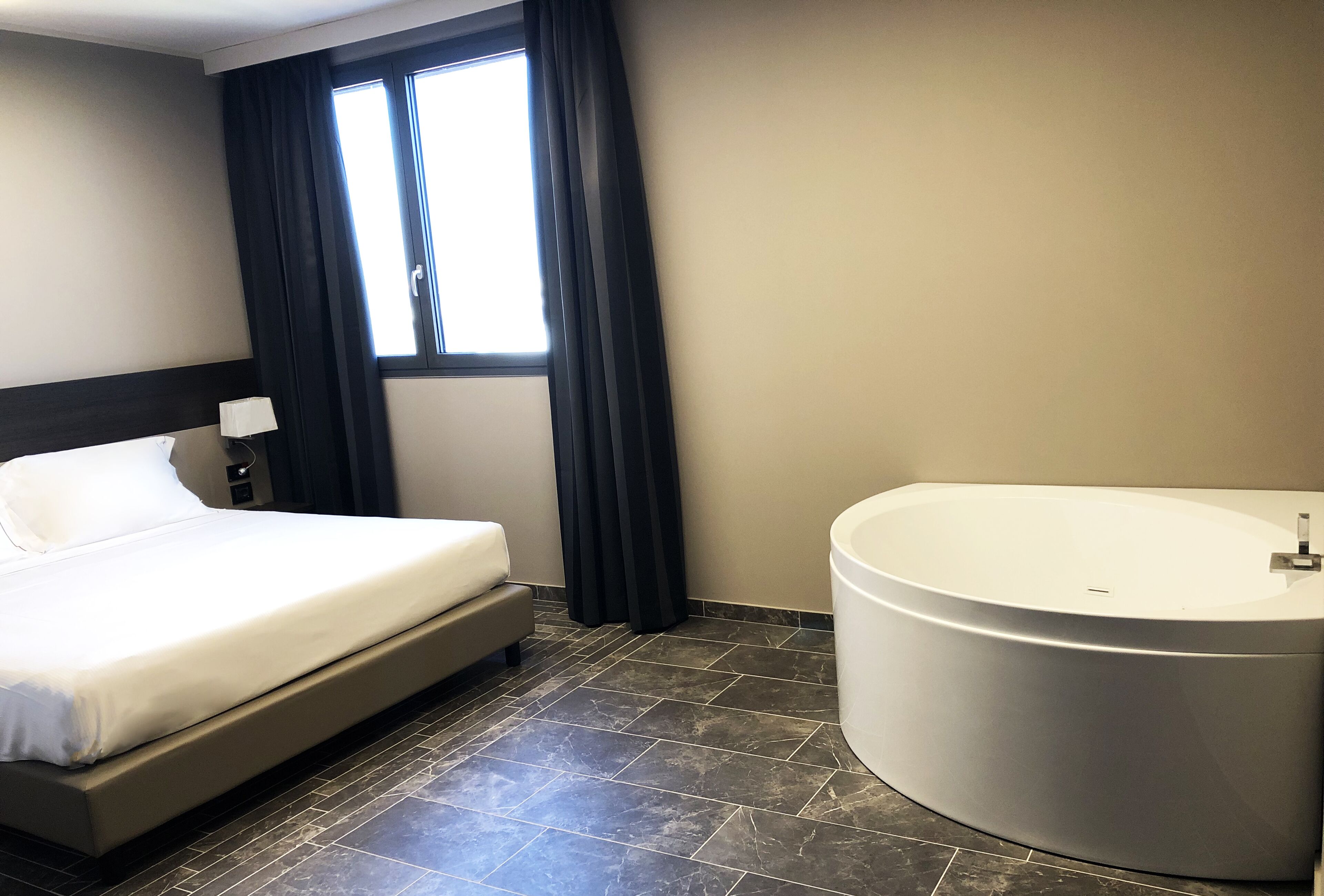 A hotel room with a bed, a large bathtub, and a window with curtains.